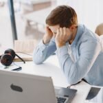 Man in a home office looks frustrated while working. He sits at a desk with a laptop and headphones.