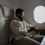 Bearded man in cozy sweater using smartphone on airplane, enjoying in-flight entertainment.